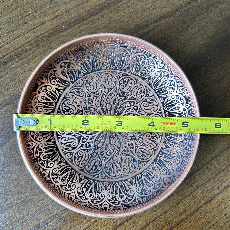 May include: A round copper-colored metal plate featuring an intricate black etched pattern of swirling lines and geometric shapes. A yellow measuring tape is placed across the center, indicating a diameter of approximately 5.5 inches.