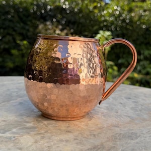 May include: A round, hammered copper mug with a polished metallic finish and a curved handle. The exterior surface features a distinct dimpled texture that reflects light, creating a warm, reddish-brown appearance across the rounded body.