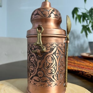 Pu&ograve; includere: Un contenitore cilindrico in rame con coperchio abbinato dotato di un pomello in ottone. Il corpo &egrave; decorato con intricati motivi incisi in nero e include una chiusura in ottone e un cucchiaio verticale in ottone fissato sul lato.
