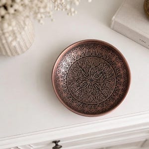 May include: A round copper-toned metal dish featuring an intricate, embossed pattern of swirling lines and symmetrical floral-inspired motifs across its interior surface, with a smooth, raised rim and a dark, oxidized finish within the recessed areas.
