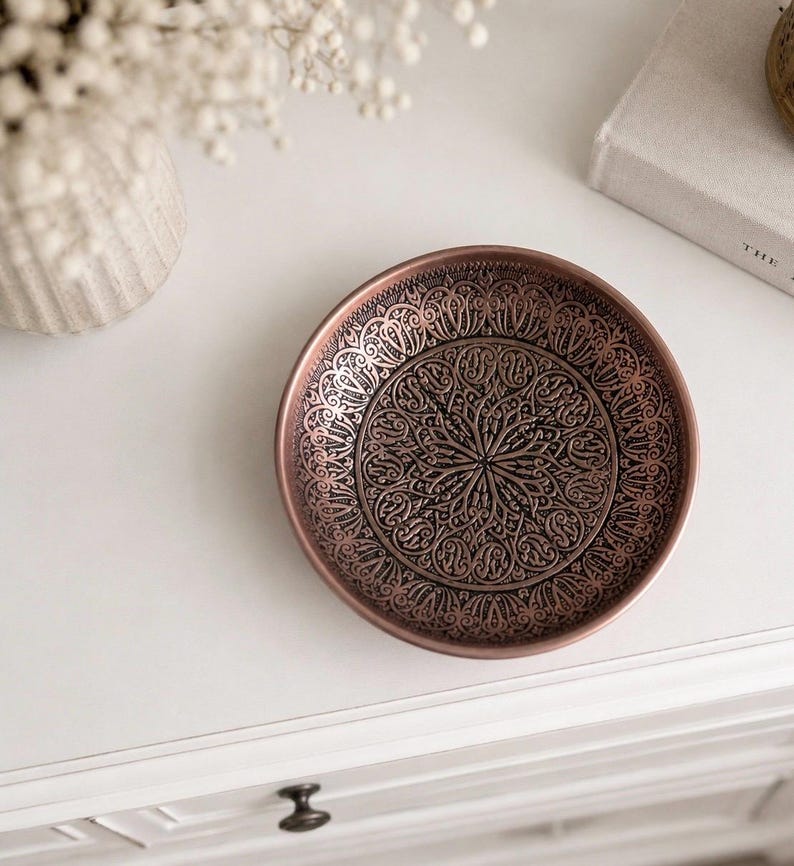 May include: A round copper-toned metal dish featuring an intricate, embossed pattern of swirling lines and symmetrical floral-inspired motifs across its interior surface, with a smooth, raised rim and a dark, oxidized finish within the recessed areas.