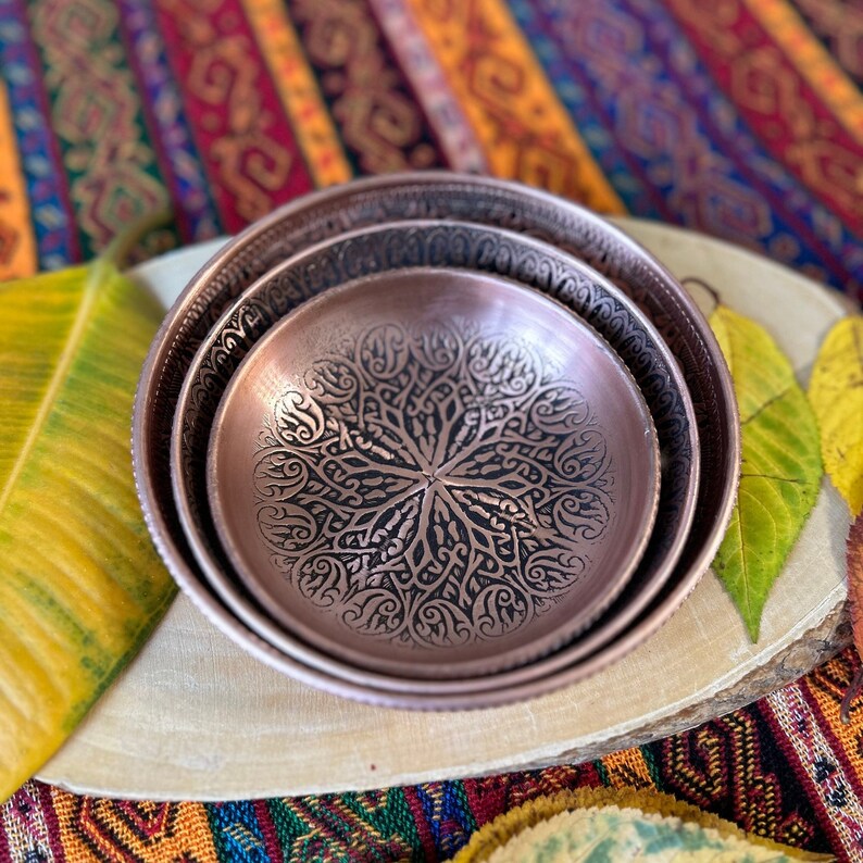 May include: Two nested circular copper bowls featuring intricate, dark, etched floral and vine patterns across their interior surfaces. The bowls have a metallic sheen and a smooth, rounded rim, showcasing a detailed, symmetrical design that radiates from the center.