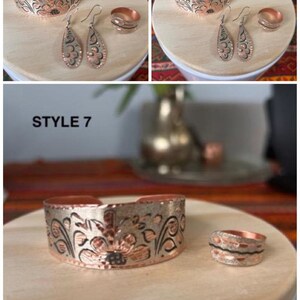 May include: A set of copper jewelry with a floral design. The set includes a bracelet, a pair of earrings, and a ring. The jewelry is made of copper and has a silver finish. The jewelry is on a wooden surface. STYLE 7