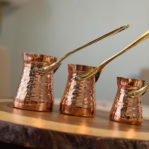 Puede incluir: Tres cafeteras de cobre con textura martillada y asas de latón. Las ollas son de diferentes tamaños, dispuestas sobre una superficie de madera. El cobre refleja la luz, creando una estética cálida. Ideal para café turco.
