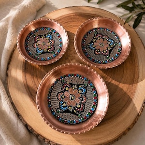 May include: Three round copper bowls with scalloped edges. Each bowl features a central black circular inlay decorated with a hand-painted floral motif in shades of pink, blue, green, and white, surrounded by a detailed gold-toned lattice pattern.