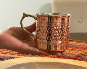 May include: A copper mug with an ornate, textured design and a brass handle. The mug has a polished finish and is held in a hand. The mug's design features a honeycomb pattern.