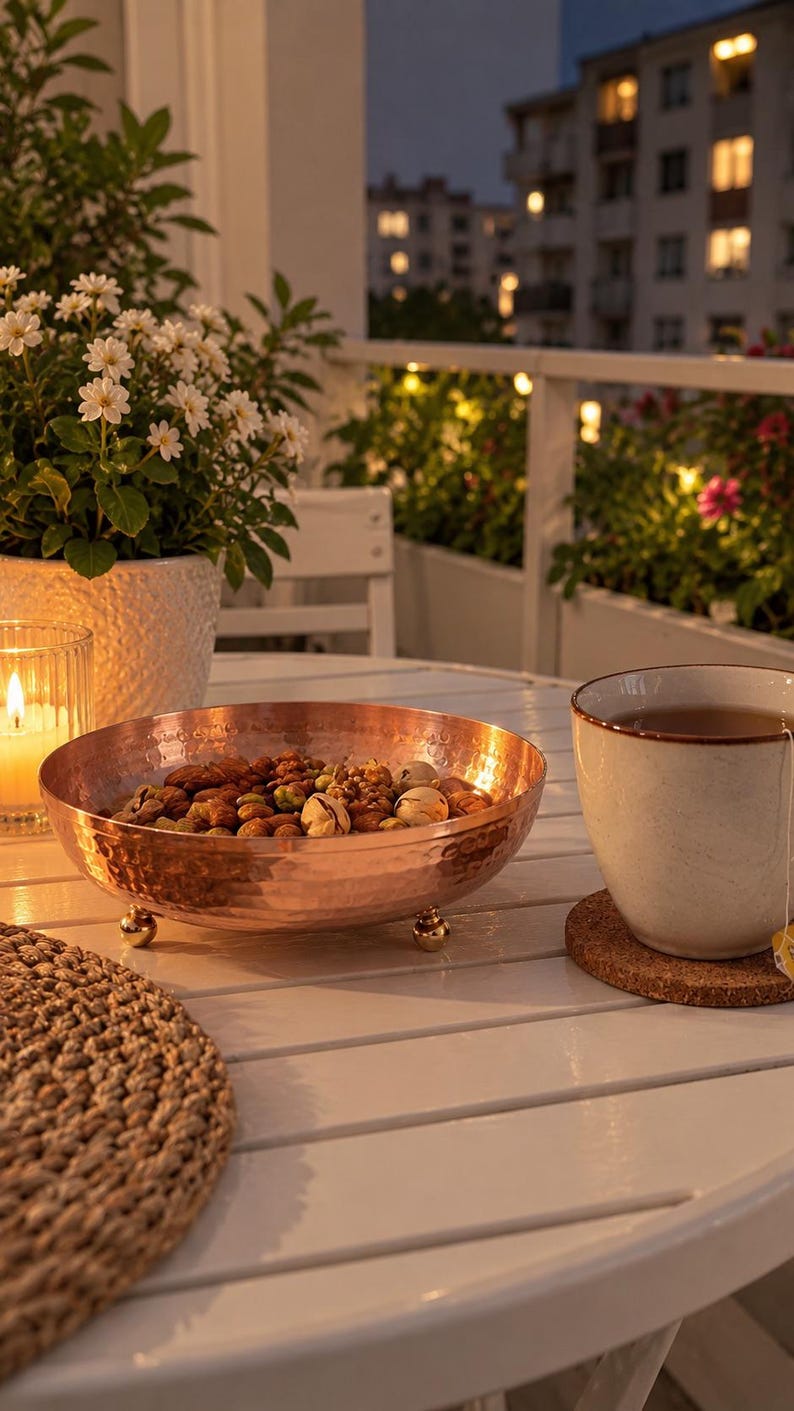 May include: A round, hammered copper bowl with a metallic finish, resting on three small, rounded gold-toned feet. The bowl is filled with a variety of mixed nuts, including almonds and pistachios, and sits on a white slatted table surface.