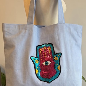 May include: Light blue tote bag with a large, sequined Hamsa hand design. The Hamsa hand features a pink center with an eye, surrounded by turquoise sequins and yellow flower accents. The bag has matching light blue handles.