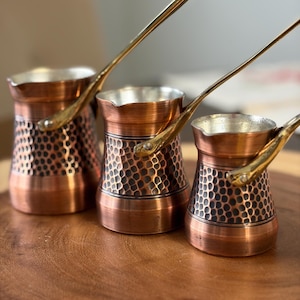 May include: Three copper coffee pots with long, gold-coloured handles. The pots have a hammered texture on the lower half and a smooth, shiny rim. They are arranged on a wooden surface, showcasing their varying sizes.