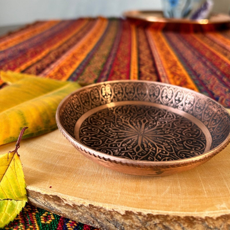 May include: A round copper dish featuring a dark, intricate, engraved floral pattern on the interior surface and a repeating heart-shaped motif along the inner rim. The exterior edge of the dish has a textured, rope-like metallic finish.