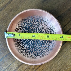 May include: A round copper bowl featuring an intricate black etched floral pattern across its interior surface. A yellow measuring tape is placed across the center, indicating a diameter of approximately 4 inches for the metallic dish.