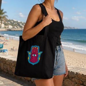 May include: A black canvas tote bag featuring a central embroidered hamsa hand design. The hamsa is rendered in vibrant shades of magenta, bright blue, and yellow, with a prominent central eye motif and small floral accents on the sides.
