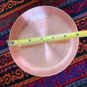 May include: A round, hammered copper bowl with a polished finish. A yellow measuring tape is placed across the center of the bowl, indicating a diameter of approximately 6.5 inches from the left edge to the right edge of the metal rim.