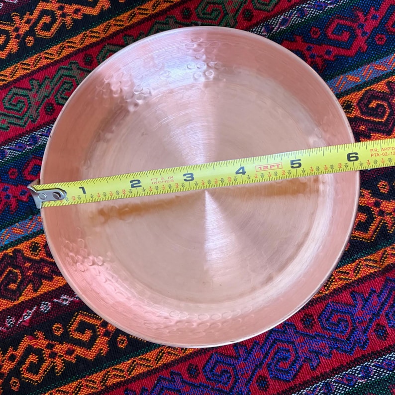May include: A round, hammered copper bowl with a polished finish. A yellow measuring tape is placed across the center of the bowl, indicating a diameter of approximately 6.5 inches from the left edge to the right edge of the metal rim.