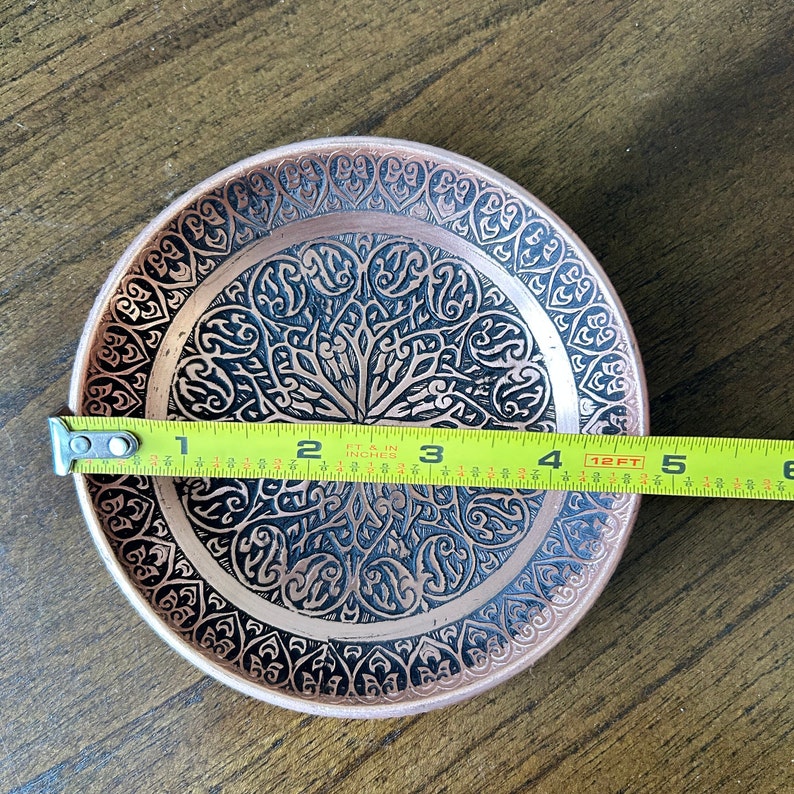 May include: A round copper-colored metal dish featuring an intricate black etched pattern of repeating floral and geometric motifs. A yellow measuring tape placed across the center shows the dish has a diameter of approximately 4.5 inches.
