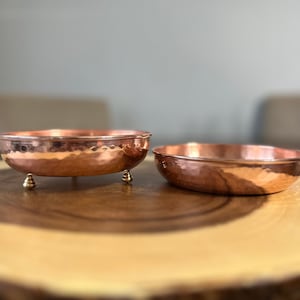 May include: Two round copper bowls with a hammered texture. The bowl on the left features three small, rounded brass-colored feet, while the bowl on the right is a simple, shallow dish without feet. Both items have a polished, metallic finish.