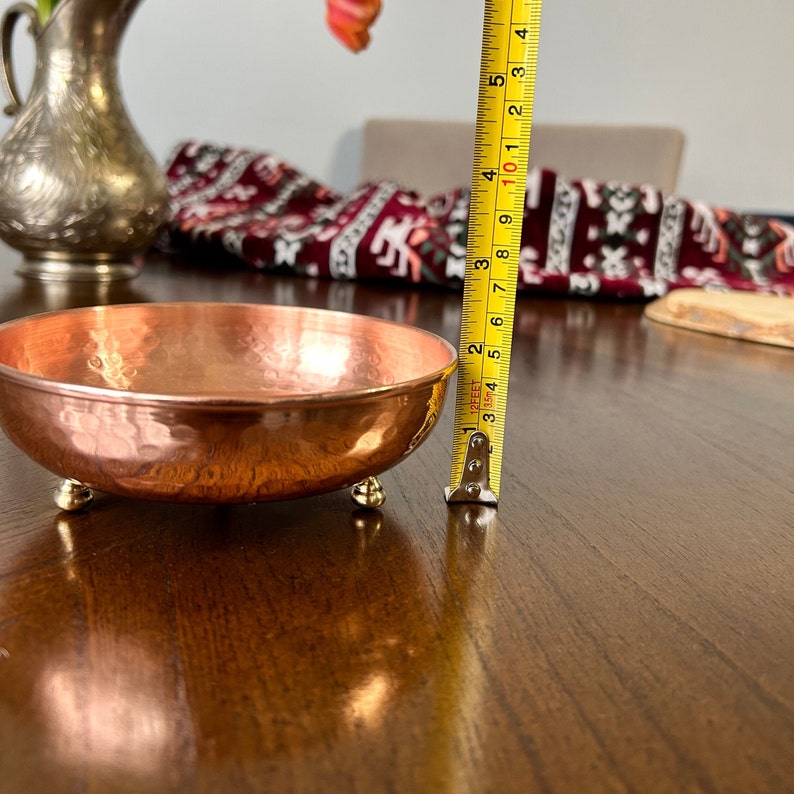 May include: A round, hammered copper bowl with three small, rounded feet. A yellow measuring tape is positioned vertically next to the bowl, indicating a height of approximately 1.5 inches for the bowl's rim from the surface.