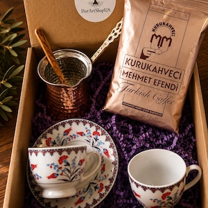 May include: A gift box containing a copper coffee pot with a wooden spoon, a 3.5 oz bag of Kurukahveci Mehmet Efendi Turkish Coffee, and two white ceramic cups with saucers featuring a red and blue floral pattern and a geometric border.