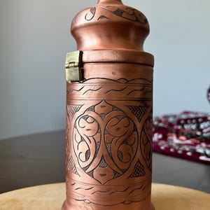Pu&ograve; includere: Un contenitore cilindrico in rame con coperchio a cupola e un piccolo pomello in ottone sulla parte superiore. Il corpo presenta intricati motivi incisi a mano e un meccanismo di chiusura in ottone sul lato con una finitura color rame.