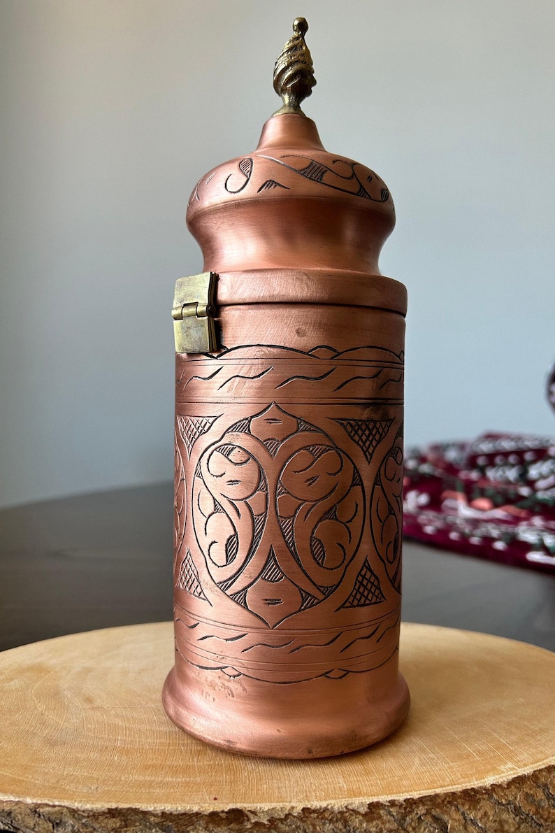 Pu&ograve; includere: Un contenitore cilindrico in rame con coperchio a cupola e un piccolo pomello in ottone sulla parte superiore. Il corpo presenta intricati motivi incisi a mano e un meccanismo di chiusura in ottone sul lato con una finitura color rame.