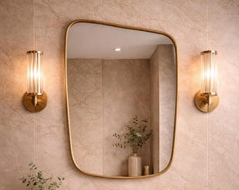 May include: A modern bathroom vanity with a gold-framed mirror, gold sconces, and a gold faucet. The vanity features a built-in sink, a tray with soap dispensers and a vase with greenery, and a stack of folded towels. The color scheme is neutral with beige and gold tones.