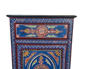May include: A small wooden cabinet featuring a single drawer and a door, painted in vibrant blue and red with intricate hand-painted geometric patterns in yellow, green, and white. The piece has a black top surface and a rectangular frame.