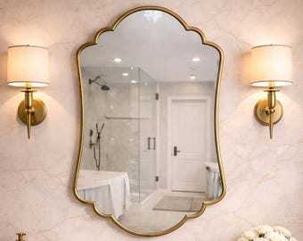 May include: A bathroom vanity with a gold-framed mirror, gold sconces, and a gold faucet. The vanity has a white sink, a marble countertop, and a gray cabinet. Decorative items include a tray with bottles and a vase of white flowers.