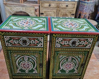May include: Two green painted wooden nightstands with intricate geometric designs. Each nightstand has a drawer and a door with a circular design. The nightstands are decorated with red, blue, yellow, and white paint.