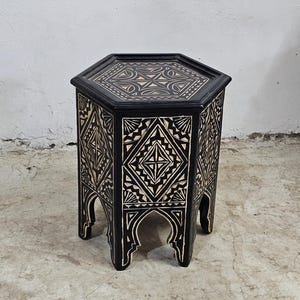 Puede incluir: Una mesa auxiliar hexagonal negra con intrincados diseños geométricos blancos tallados en la madera. La mesa tiene un diseño simple y elegante y sería una gran adición a cualquier hogar.