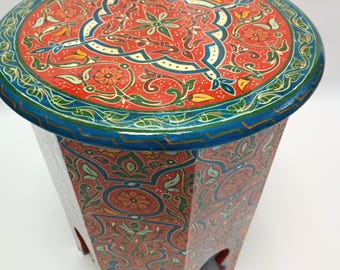 May include: A round, hand-painted wooden side table with a vibrant, intricate design. The tabletop features a red background with a detailed floral and geometric pattern in blue, green, and yellow. The sides are also decorated with matching patterns. The table is approximately 18 inches in diameter and 18 inches tall.