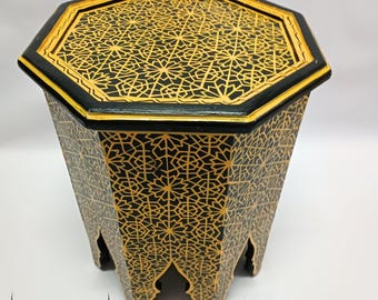 May include: A black and gold octagonal side table with intricate geometric patterns. The table has a black base and a gold-colored top with a decorative border. The table is approximately 20 inches tall.