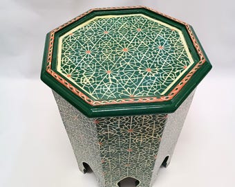 May include: Green and cream-colored octagonal side table with a geometric pattern. The table has a dark green border with orange accents. The legs have a decorative cutout design. A decorative accent piece for any room.