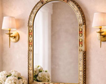 May include: An arched mirror with a decorative gold frame featuring red and yellow accents. The mirror is flanked by two sconces with white lampshades. A bouquet of white flowers sits below the mirror, adding a touch of elegance.