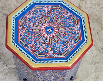 May include: Octagonal wooden table with intricate geometric patterns painted in blue, red, gold, and white. The table has a unique, carved base.