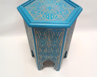 May include: A teal hexagonal side table with gold floral carvings. The top features a large floral design, and the sides have repeating geometric patterns. The table has arched cutouts at the base and is made of wood.