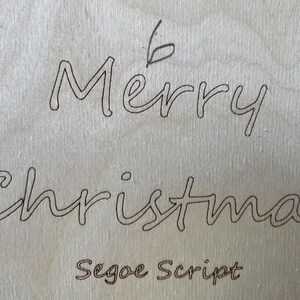 May include: A close-up of a wooden sign with the words "Merry Christmas" written in cursive script. The text "Segoe Script" is written below the greeting.