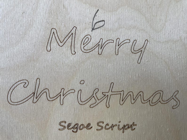 May include: A close-up of a wooden sign with the words "Merry Christmas" written in cursive script. The text "Segoe Script" is written below the greeting.