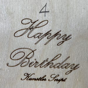 May include: A wooden sign with the words "Happy Birthday" written in cursive script. The number "4" is written above the greeting. The text "Kunster Script" is written below the greeting.