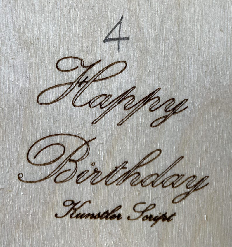 May include: A wooden sign with the words "Happy Birthday" written in cursive script. The number "4" is written above the greeting. The text "Kunster Script" is written below the greeting.