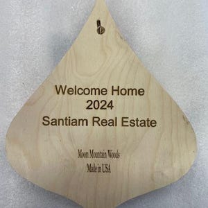 May include: A wooden, teardrop-shaped ornament with the text "Welcome Home 2024 Santiam Real Estate Moon Mountain Woods Made in USA".