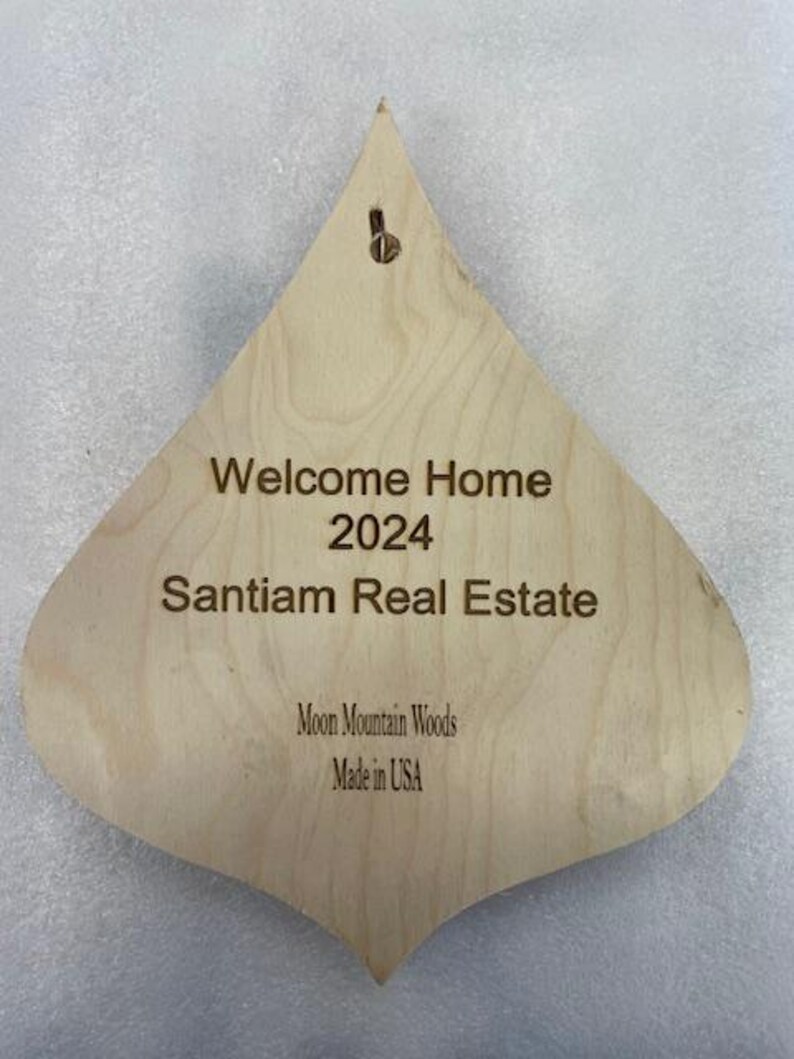May include: A wooden, teardrop-shaped ornament with the text "Welcome Home 2024 Santiam Real Estate Moon Mountain Woods Made in USA".