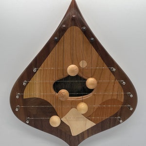 May include: A teardrop-shaped wooden musical instrument with a dark wood frame and a light wood center. The instrument has strings and small wooden balls that hang from the strings.