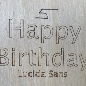 May include: A wooden sign with the text "Happy Birthday" in a black outline font. The font is identified as "Lucida Sans".