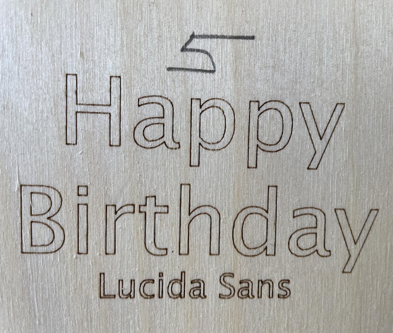 May include: A wooden sign with the text "Happy Birthday" in a black outline font. The font is identified as "Lucida Sans".
