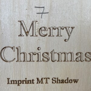 May include: A wooden sign with the text "Merry Christmas" and the words "Imprint MT Shadow" below. The sign is a light brown color and has a slightly rough texture.