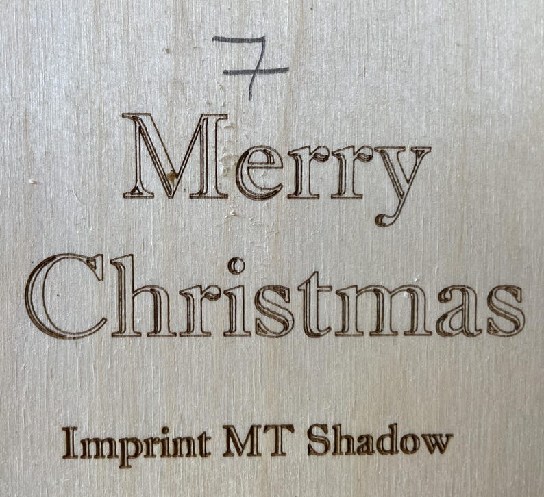 May include: A wooden sign with the text "Merry Christmas" and the words "Imprint MT Shadow" below. The sign is a light brown color and has a slightly rough texture.