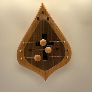 Può includere: Uno strumento musicale in legno a forma di lacrima con una croce tagliata al centro. Lo strumento ha tre corde e tre pomelli in legno che vengono utilizzati per suonare lo strumento.