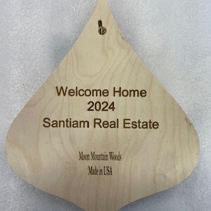 May include: A wooden, teardrop-shaped ornament with the text "Welcome Home 2024 Santiam Real Estate Moon Mountain Woods Made in USA".