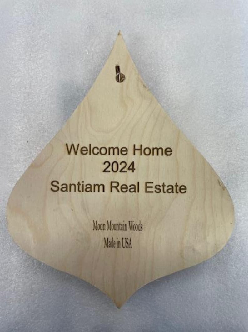 May include: A wooden, teardrop-shaped ornament with the text "Welcome Home 2024 Santiam Real Estate Moon Mountain Woods Made in USA".