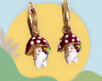 May include: Gold hoop earrings with a red and white mushroom charm. The mushroom has a white face with a smile and is holding a bouquet of colorful flowers.